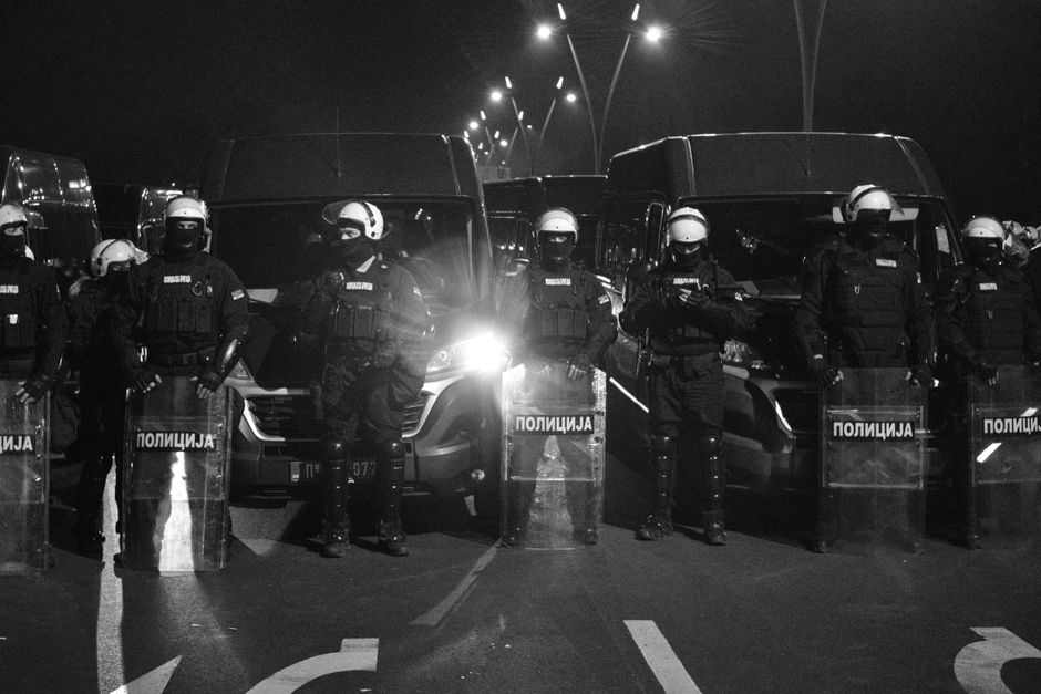 Riot police in protective gear and shields stand in line at night.